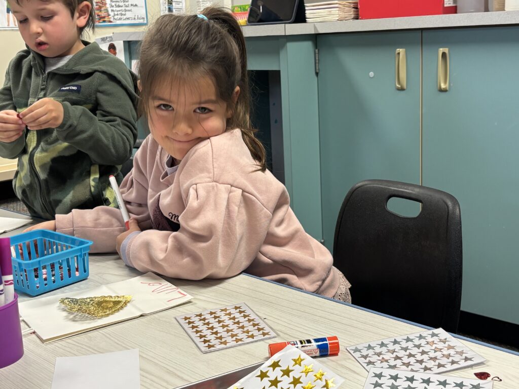 A young girl with long hair smiles while working on a craft project, surrounded by stickers and art supplies.