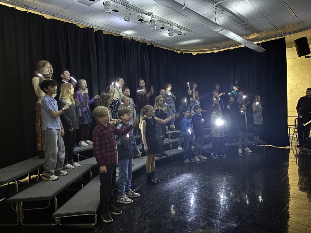 A group of children performing on stage, holding flashlights under bright lights with a black curtain backdrop.