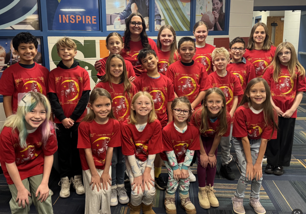 A group of children wearing red Lunar New Year t-shirts smiles for a photo in a school hallway.