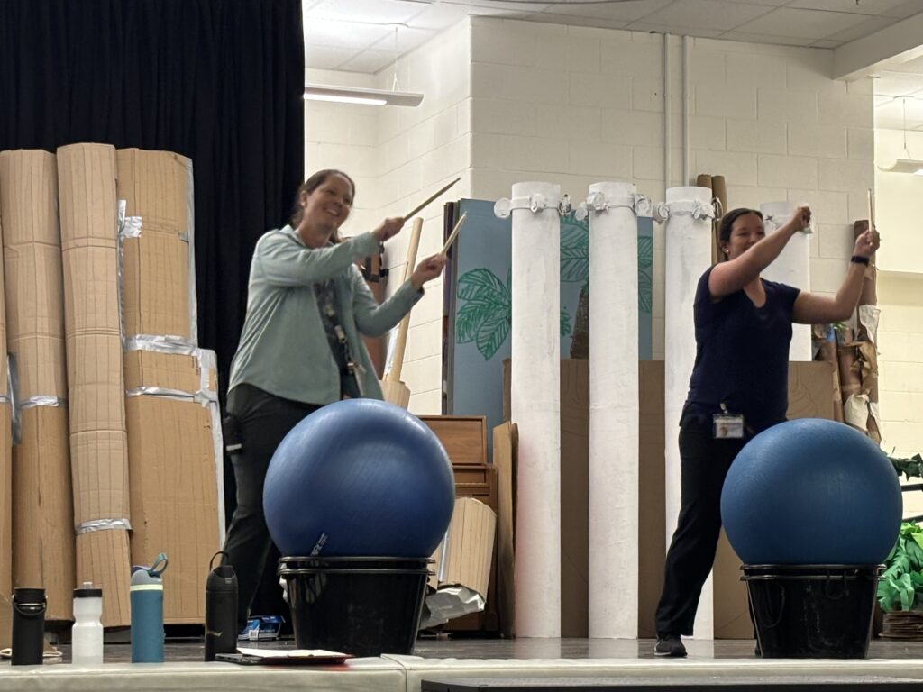 Two women are performing on stage with blue exercise balls and buckets, surrounded by cardboard props.