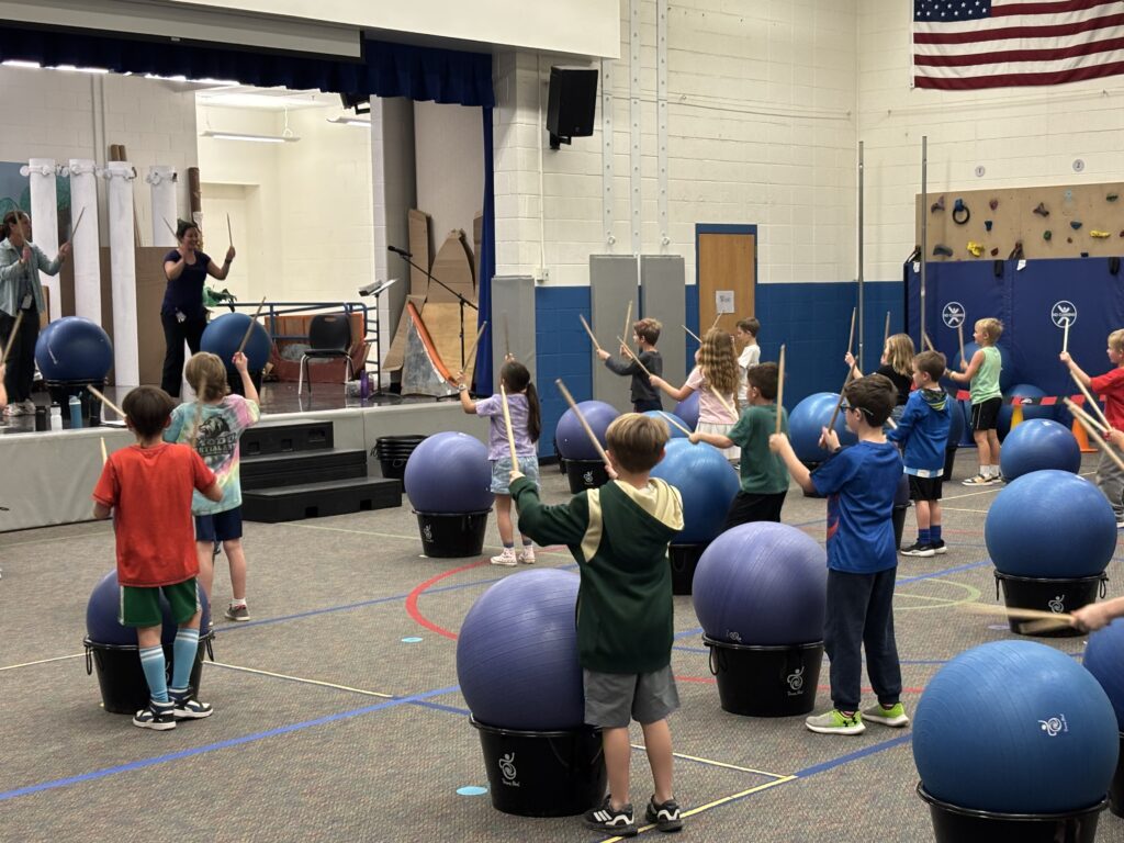 Children participate in a rhythmic drumming session using exercise balls and drumsticks in a gym setting.
