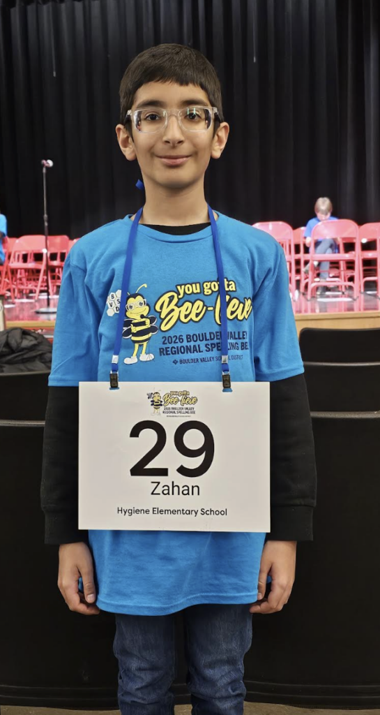 A boy wearing a blue t-shirt and glasses stands with a sign reading "29 Zahan, Hygiene Elementary School" in a gym.