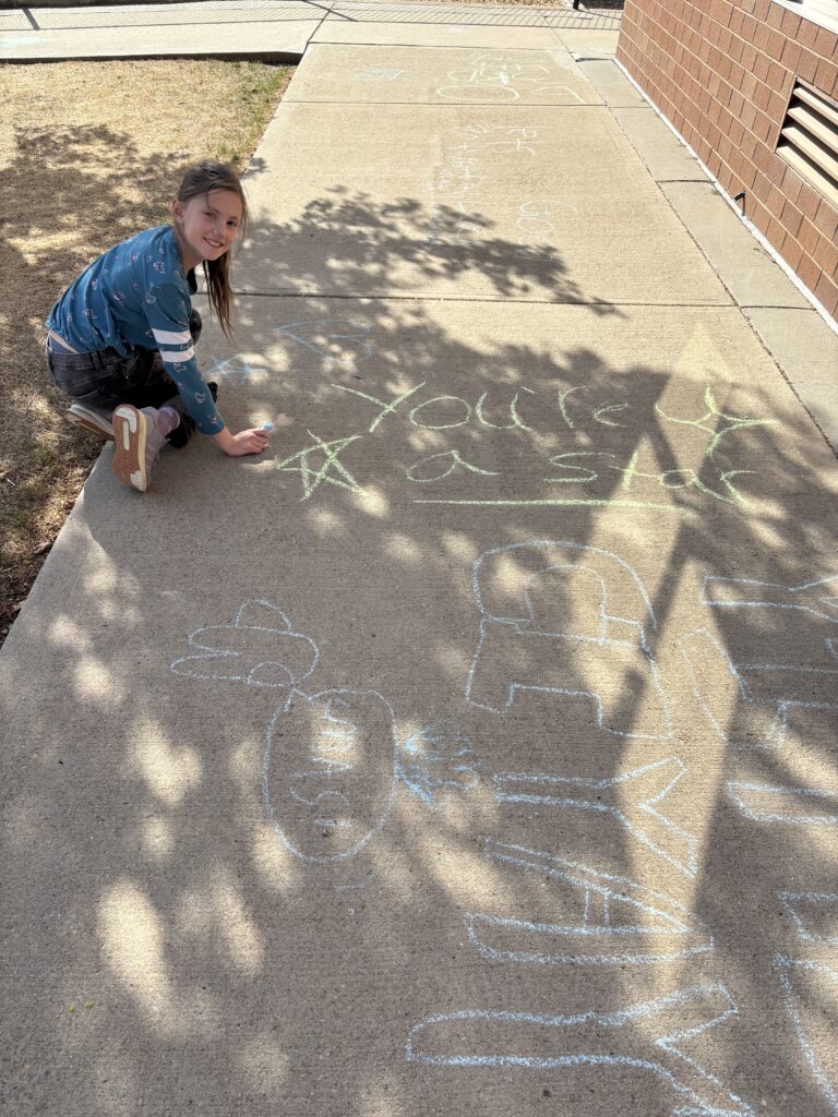 A child kneels on a sidewalk, drawing with chalk, while writing "You're a star" surrounded by colorful doodles.
