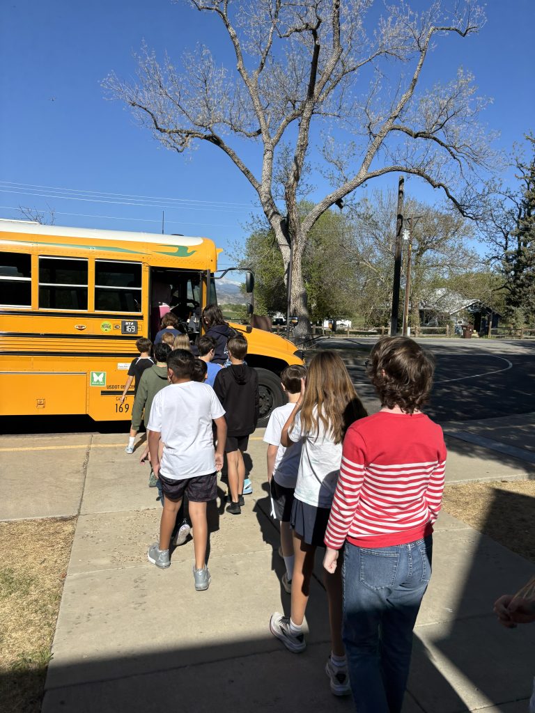 A line of children boarding a yellow school bus on a sunny day, with a clear blue sky and bare trees in the background.