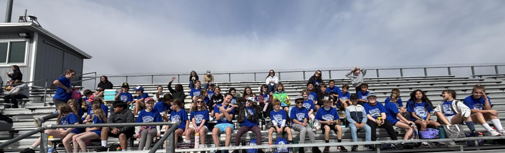 Un numeroso grupo de niños con camisetas azules sentados en las gradas, sonriendo y disfrutando de un día soleado en un evento al aire libre.