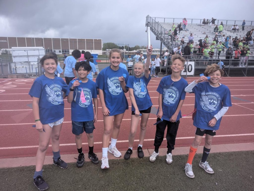 Un grupo de seis niños con camisetas azules está de pie en una pista, sonriendo y sosteniendo cintas, con espectadores al fondo.
